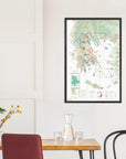 A De Long Wine Map of Greece Framed hangs on a white wall above a dining table set with glasses, carafe, and plant.
