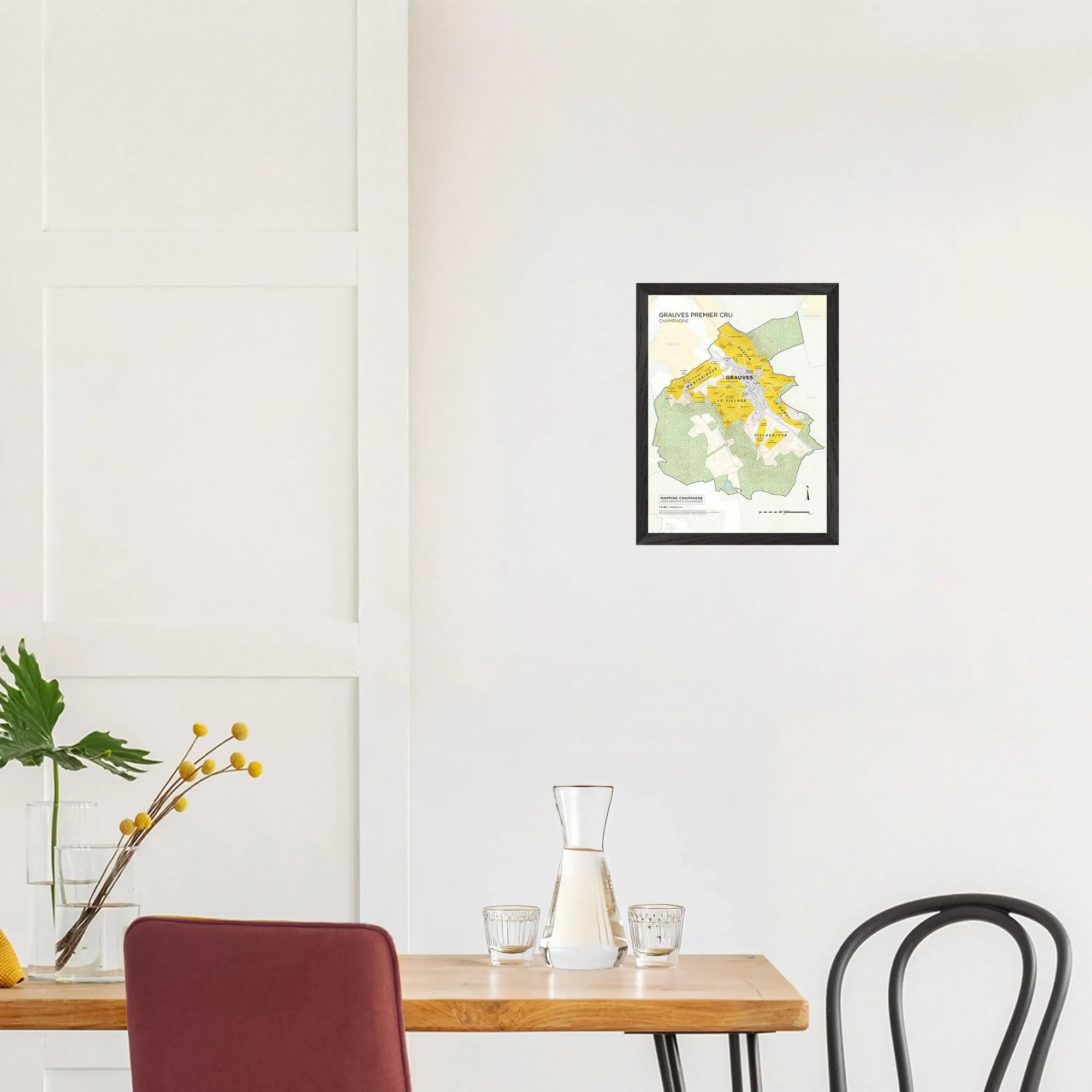 Minimal dining area with small table, chairs, glassware, and De Long Grauves Premier Cru Côte des Blancs Framed on white wall.