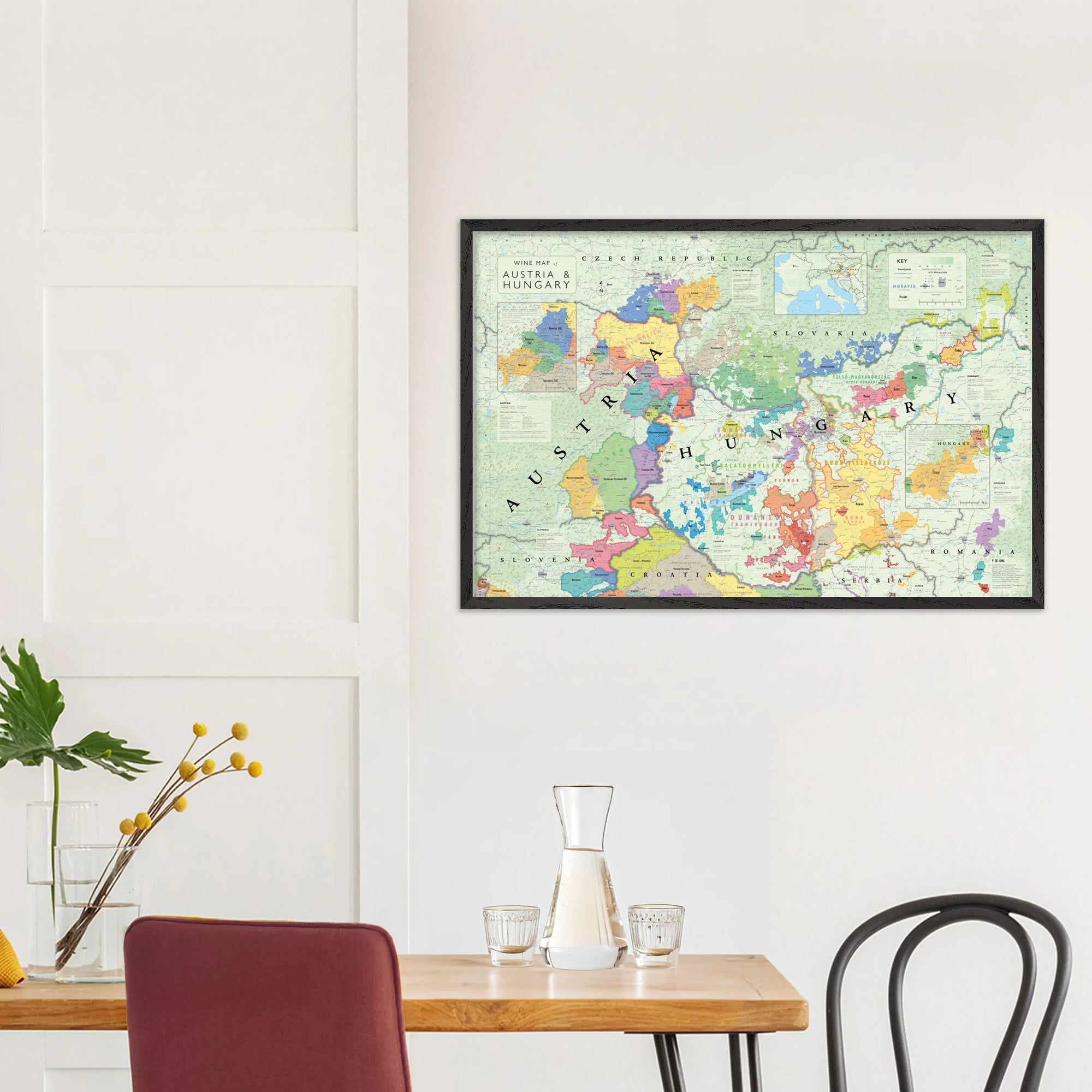 The De Long Wine Map of Austria and Hungary Framed hangs on a white wall above a small dining table with chairs.