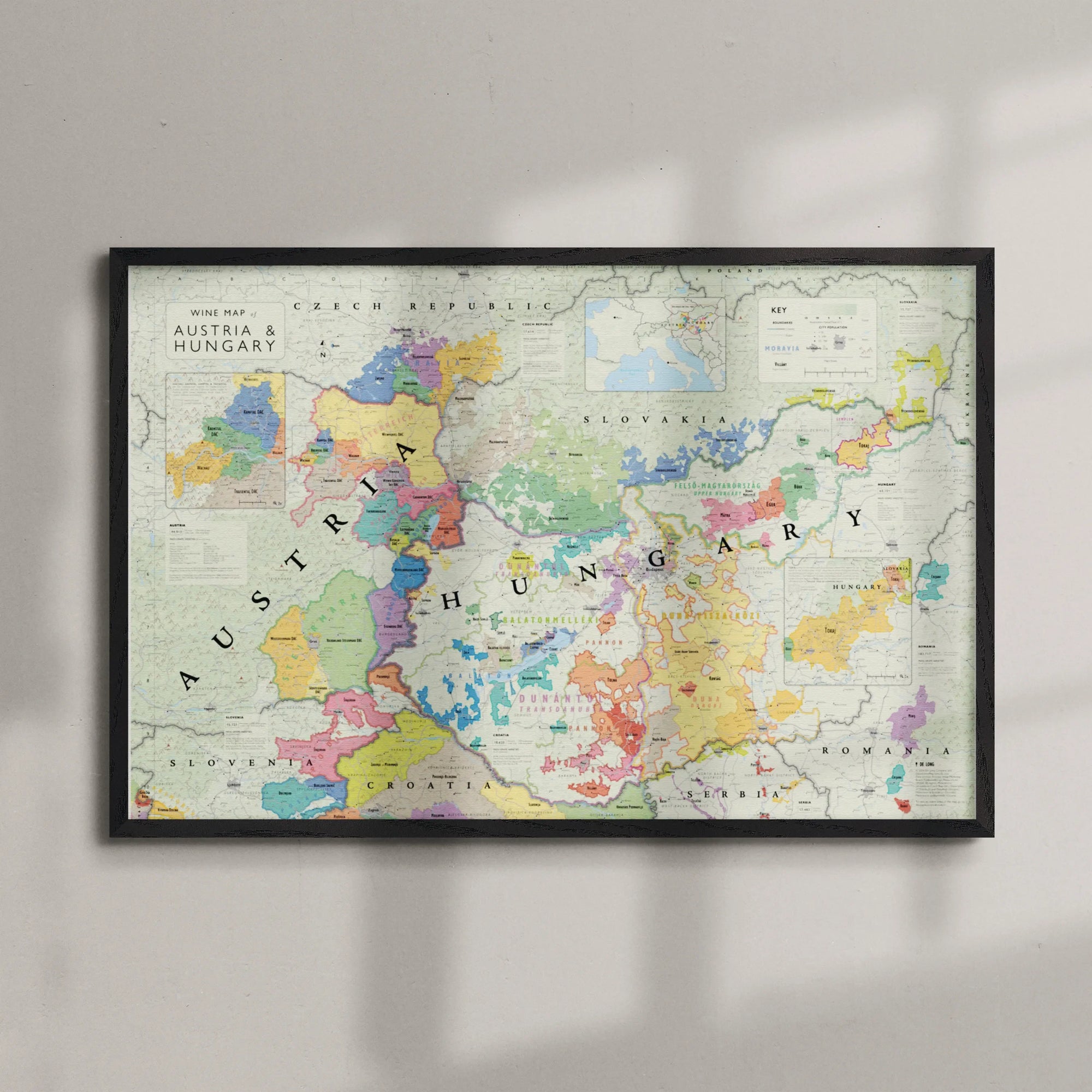 The Wine Map of Austria and Hungary Framed features vibrant regional boundaries and labels, displayed on a light-colored wall with soft shadows enhancing its elegant presentation.