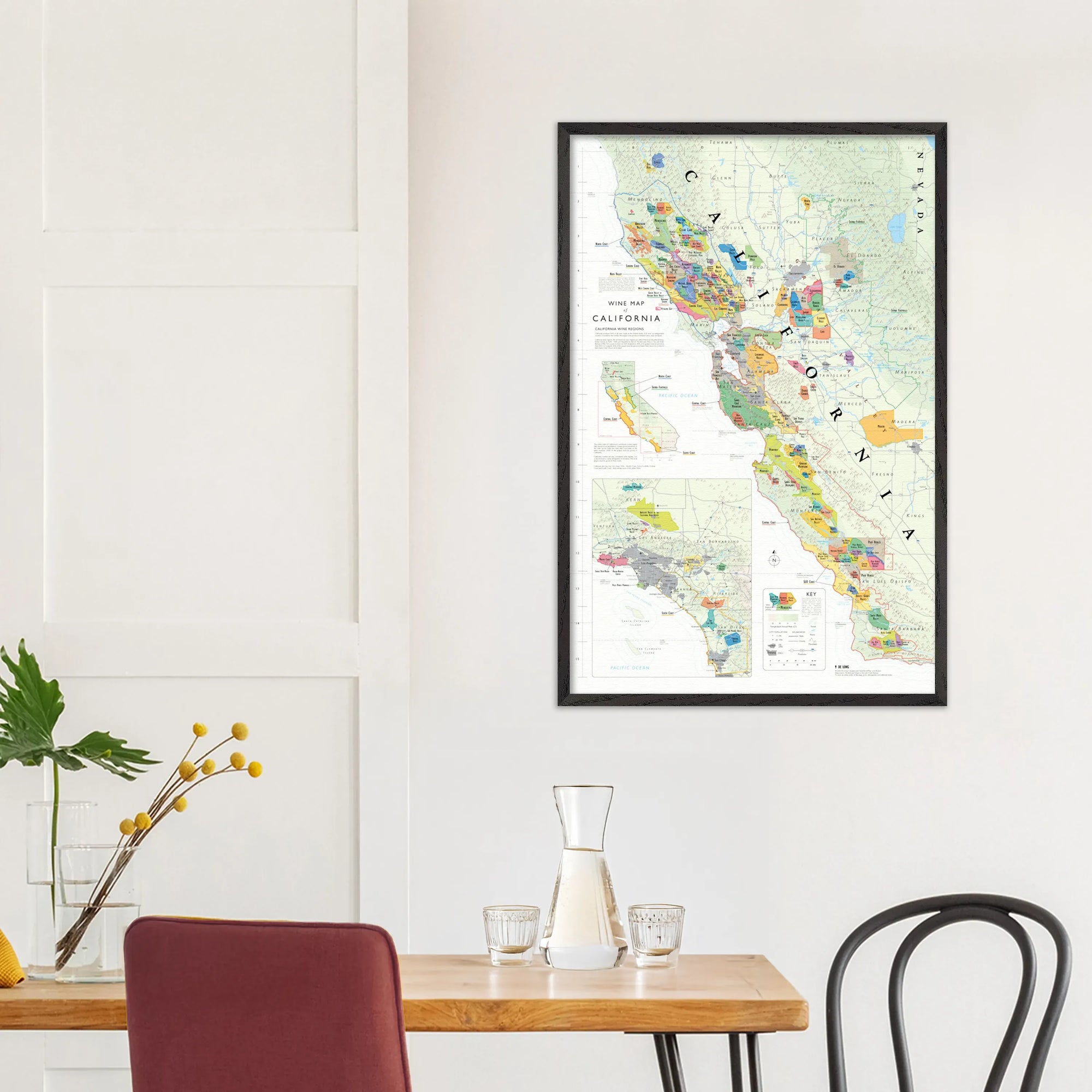 De Long Wine Map of California Framed (24x36") showcased above a dining table with chairs, glassware, and a plant.