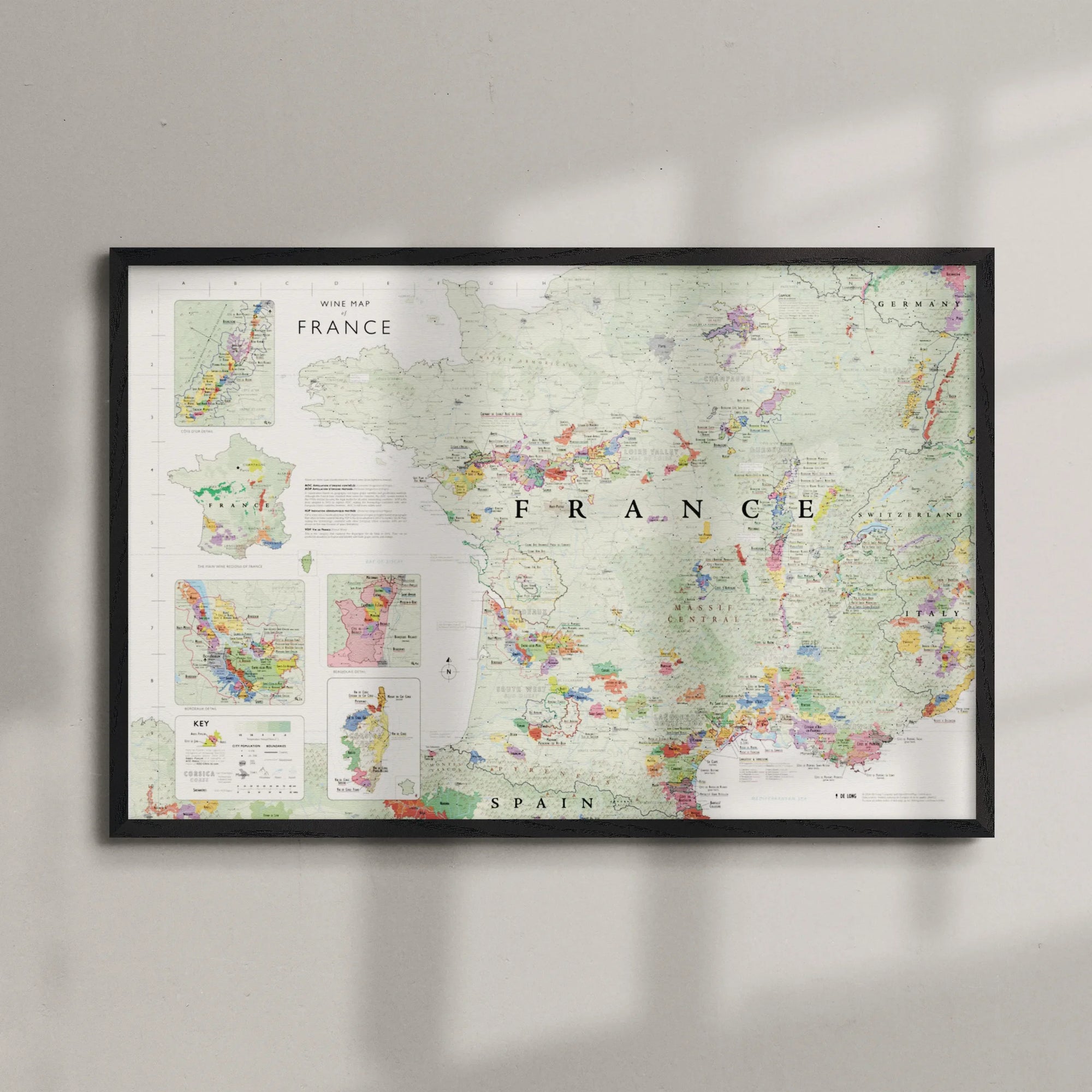 The Wine Map of France Framed features colored and labeled wine regions, highlighting detailed geography. Displayed in a wood frame, it hangs on a light-colored wall with gentle shadows creating an elegant effect.