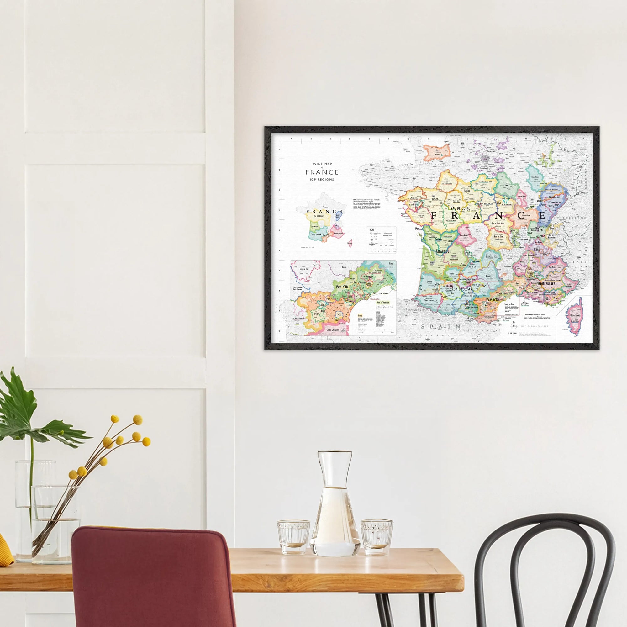 The De Long Wine Map of France IGPs Framed hangs on a white wall above a minimalist dining table.