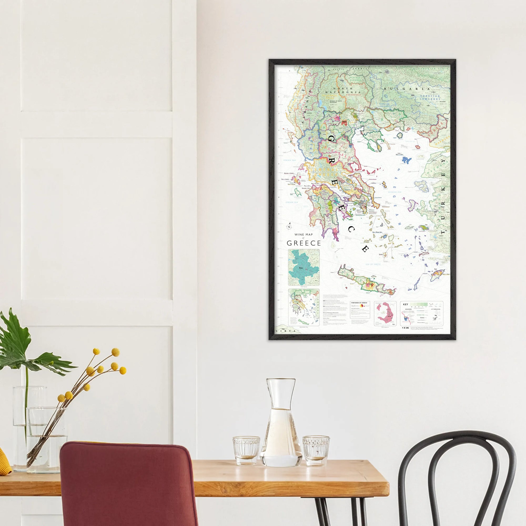 A De Long Wine Map of Greece Framed hangs on a white wall above a dining table set with glasses, carafe, and plant.