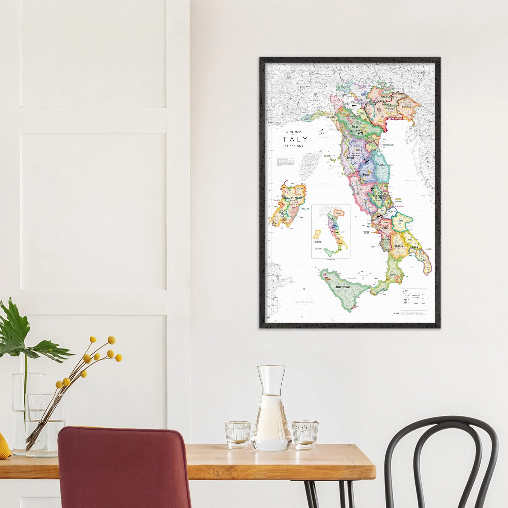 A De Long Wine Map of Italy IGTs Framed hangs on a white wall above a dining table with chairs and a glass carafe.