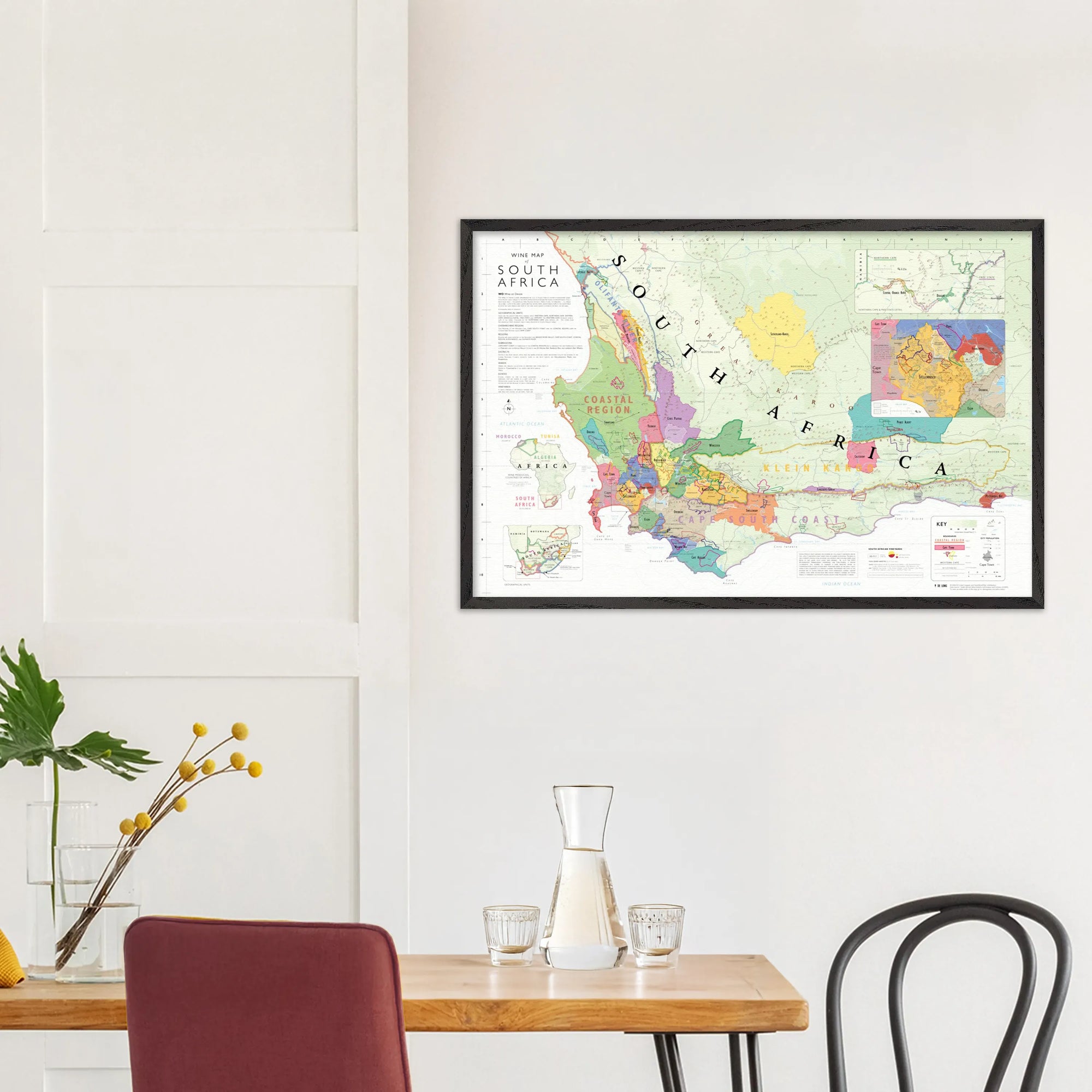 The De Long Wine Map of South Africa Framed hangs above a small dining table with two chairs and a glass carafe.