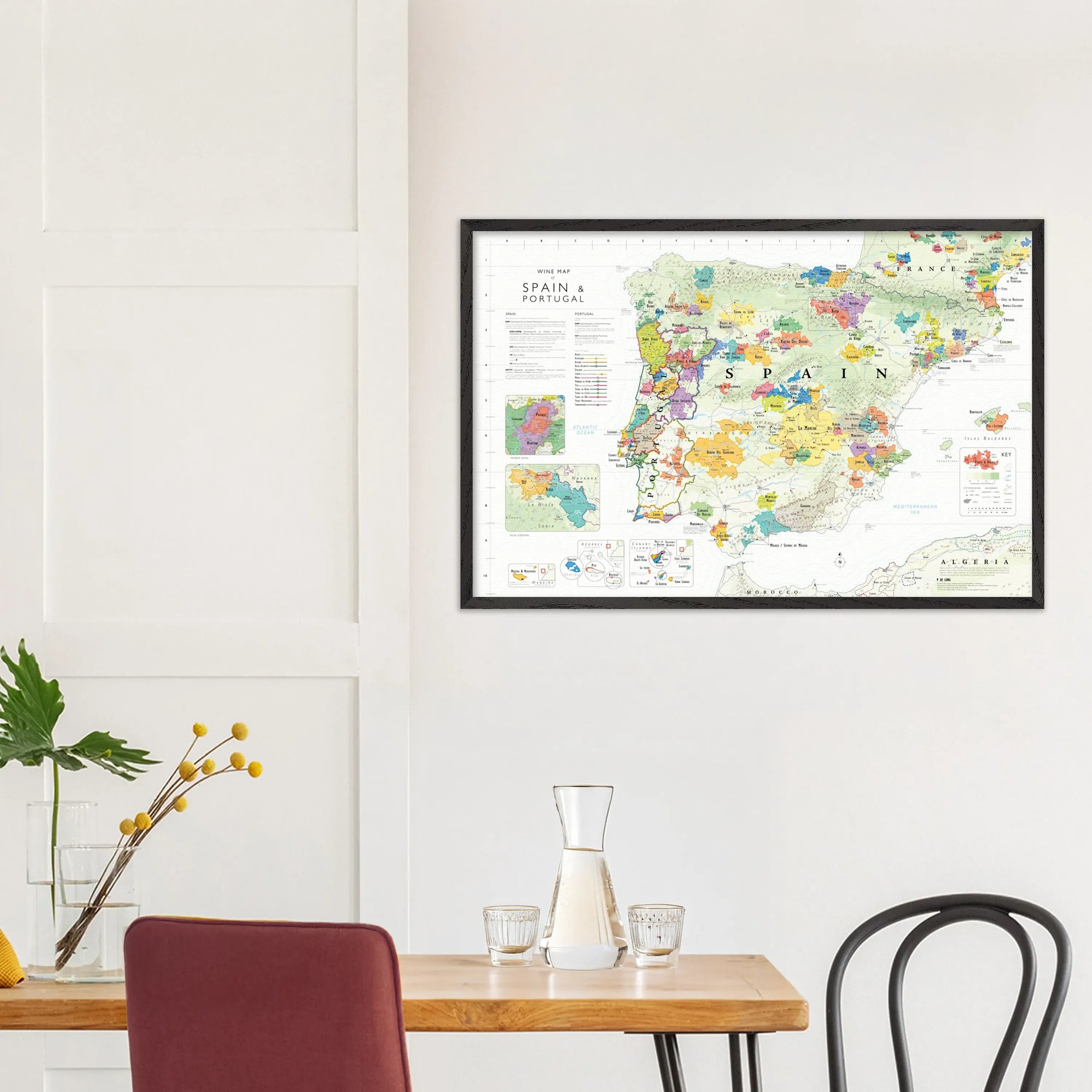 A De Long Wine Map of Spain &amp; Portugal Framed hangs above a dining table with chairs and a glass carafe.