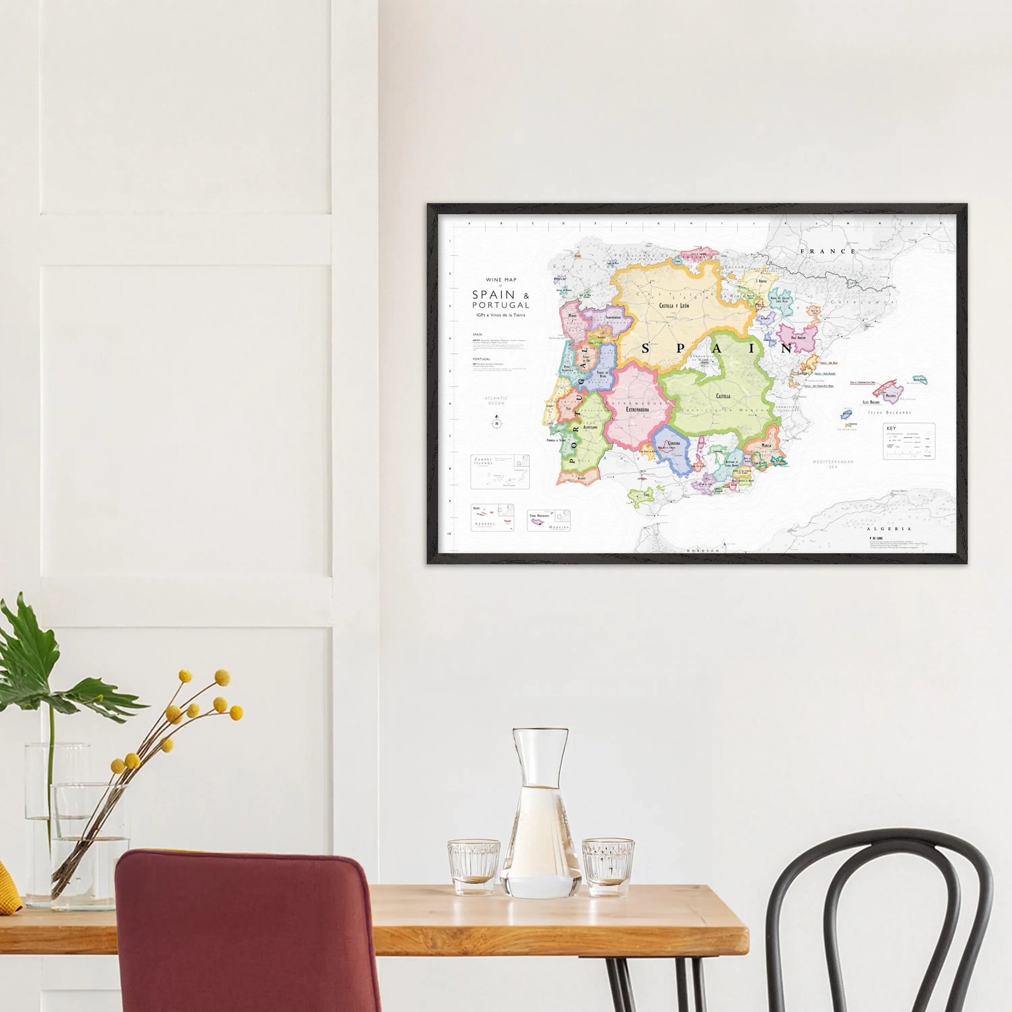 A De Long Wine Map of Spain &amp; Portugal IGPs Framed hangs above a wooden table with a carafe, glasses, and a plant.