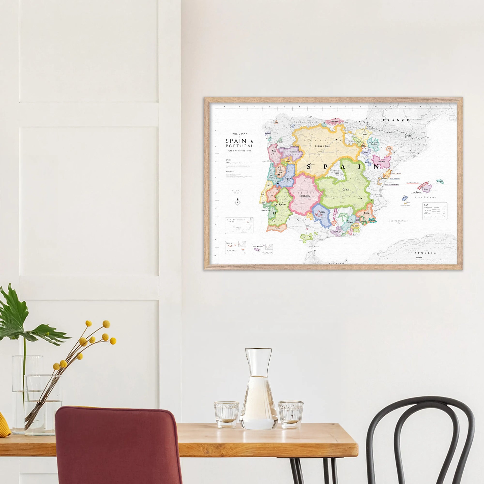 A De Long Wine Map of Spain &amp; Portugal IGPs, framed, hangs above a table with carafe, glass, vase, and chair.