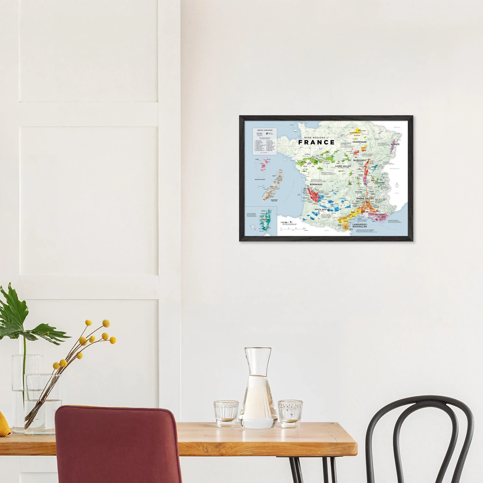 A De Long Wine Regions of France Vino One Framed map hangs above a dining table with chairs, carafe, and yellow flowers.
