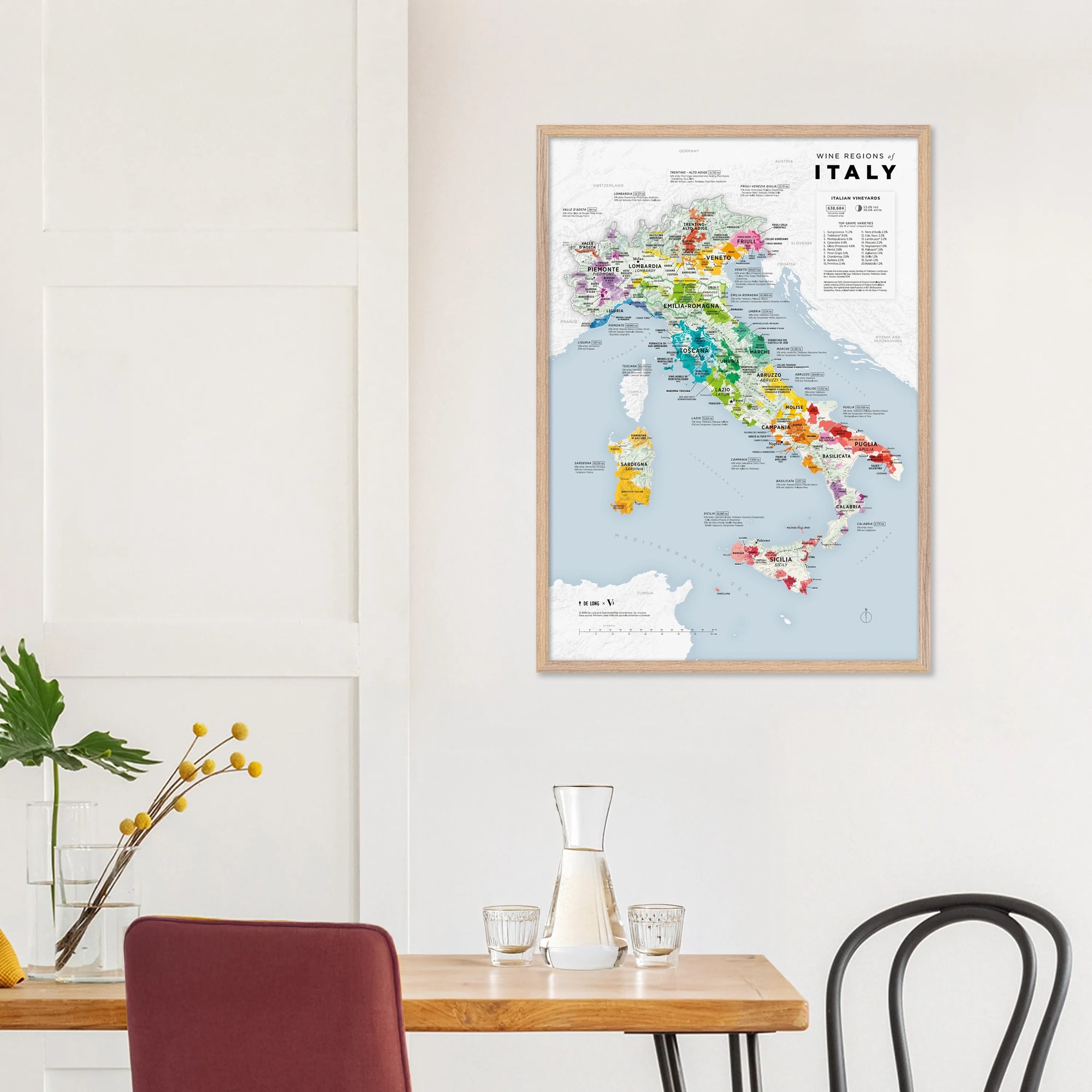 A De Long Wine Regions of Italy Vino One Framed map hangs above a dining table with chairs, a vase, and glassware.