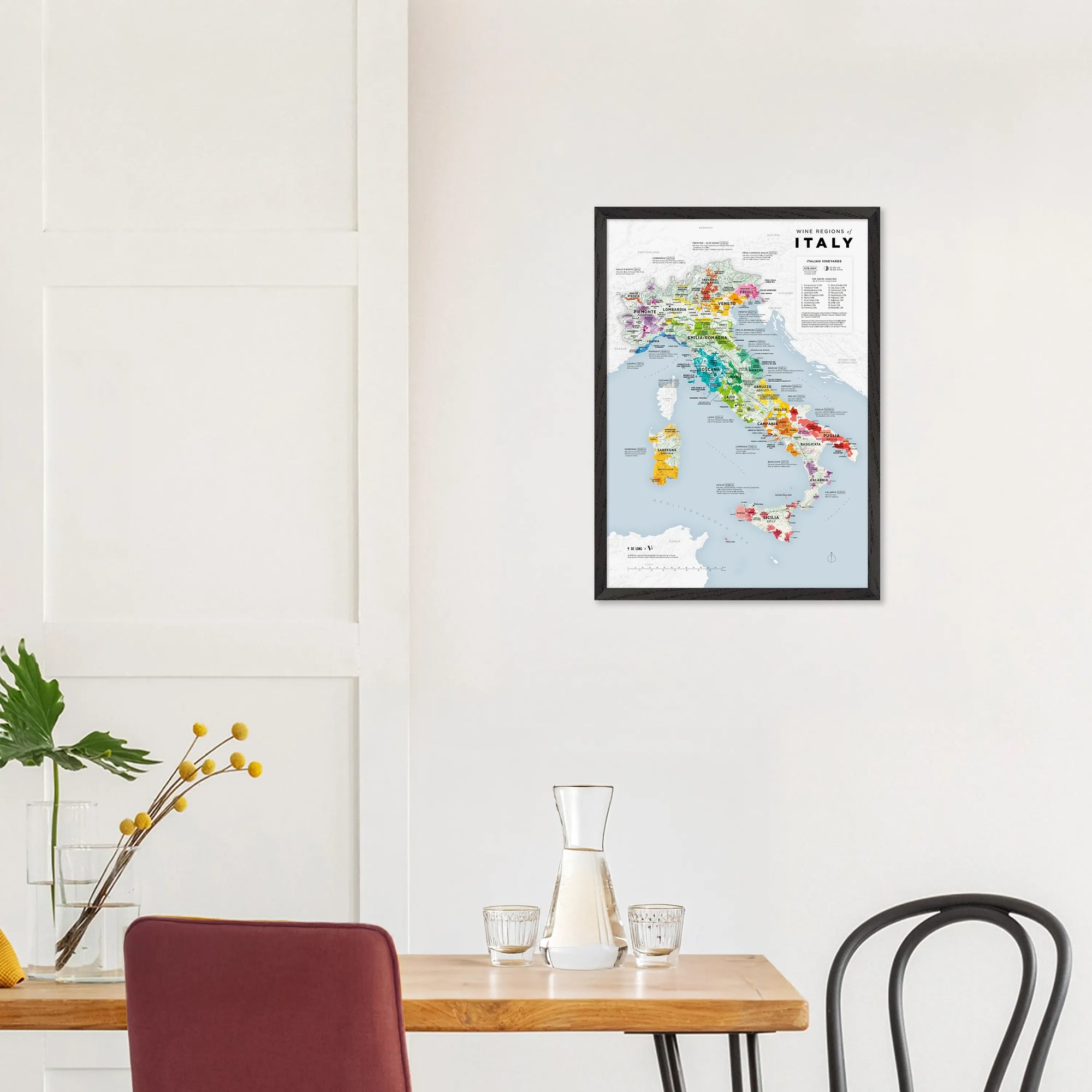 The De Long Wine Regions of Italy Vino One Framed map hangs on a white wall above a table with chairs and decor.