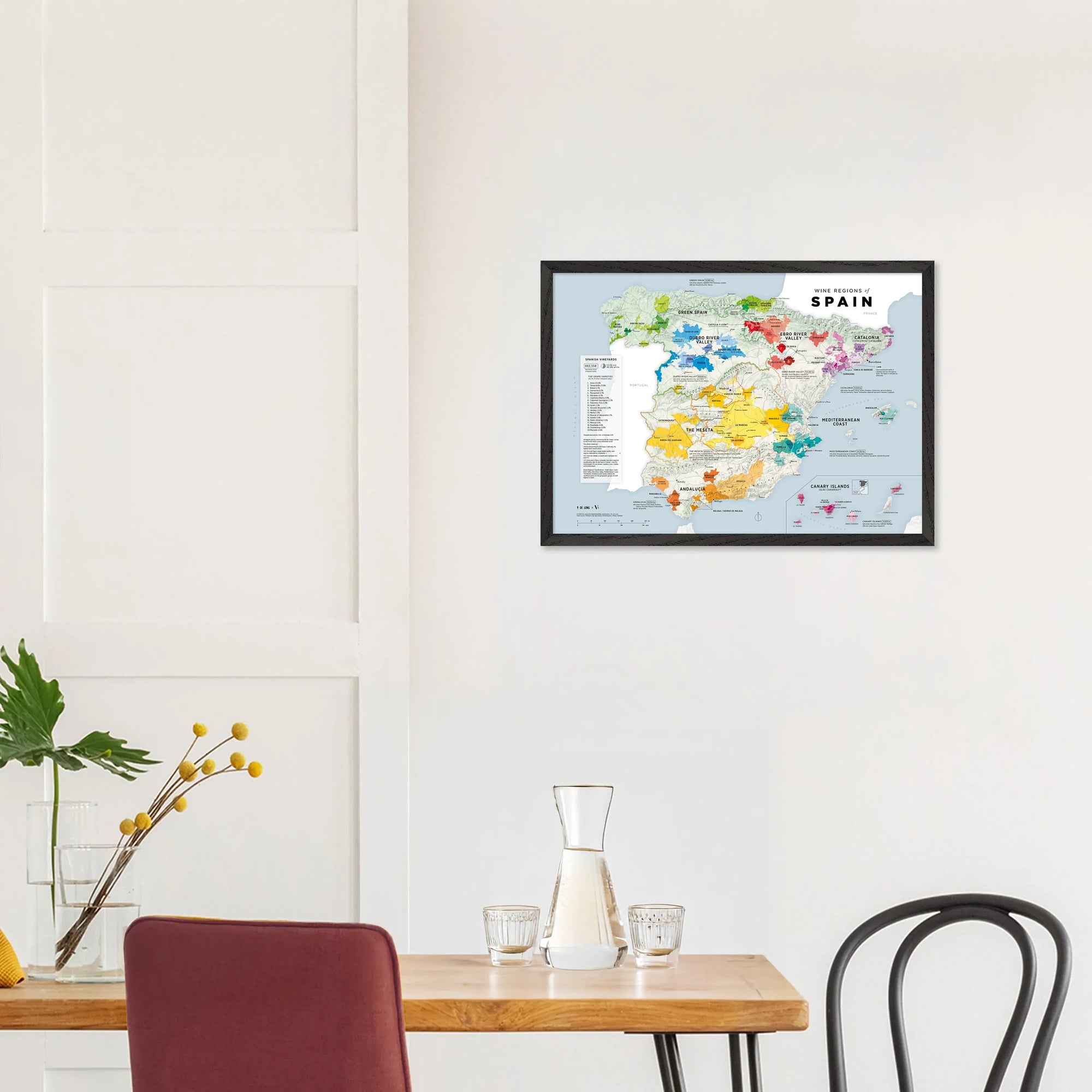 A De Long Wine Regions of Spain Vino One Framed map hangs above a dining table with chairs and a small plant.